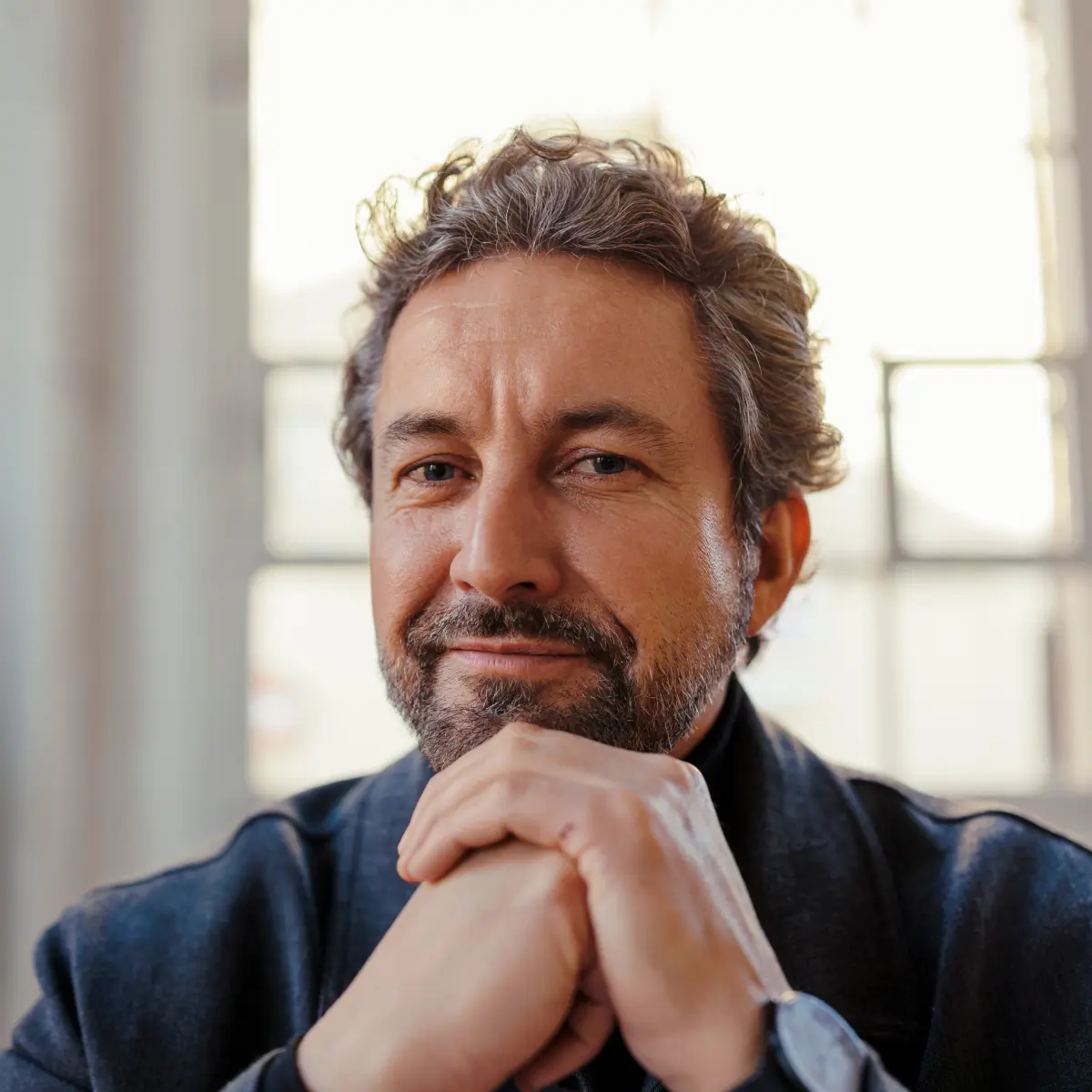 Lächelnder Mann mit Bart und graumeliertem Haar stützt sein Kinn auf die gefalteten Hände.