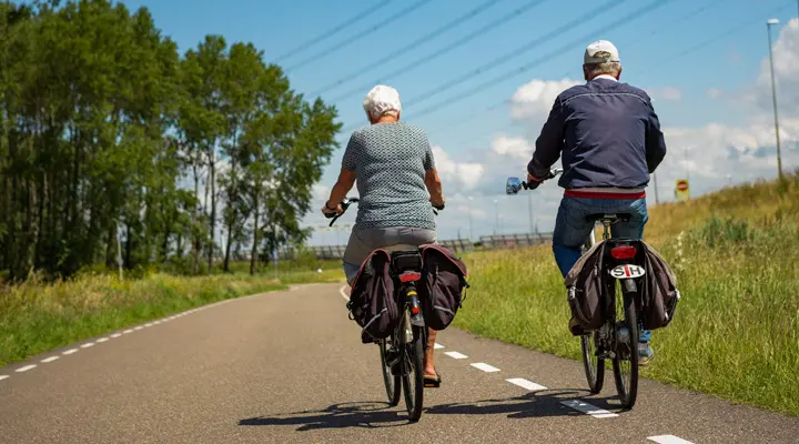 Twee personen fietsen over een asfaltweg tussen groene velden in een landelijke omgeving.