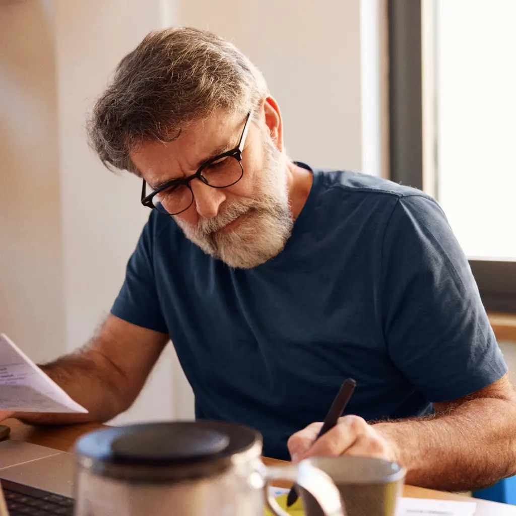 A mature man with glasses and a grey beard wearing a blue t-shirt sits at a desk, reviewing paperwork and taking notes with a pen.