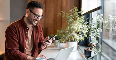 raisin_fi_bank_switch.jpg Mies hymyilee ja katsoo puhelinta tietokoneen ääressä.