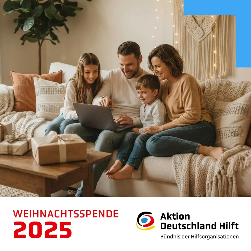 Eine vierköpfige Familie sitzt gemütlich auf einem Sofa und schaut gemeinsam auf einen Laptop. Im Vordergrund stehen drei brennende Kerzen auf einem Holztisch. Unten im Bild befindet sich ein weißer Bereich mit roter Aufschrift ‚Weihnachtsspende 2025‘ sowie dem Logo von ‚Aktion Deutschland Hilft – Bündnis der Hilfsorganisationen‘.