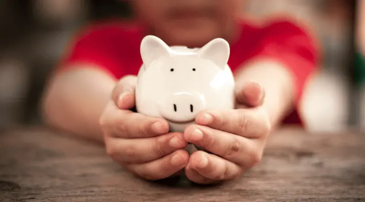 Dos manos cogen una hucha blanca con forma de cerdo. La persona que coge la hucha lleva una camiseta roja.