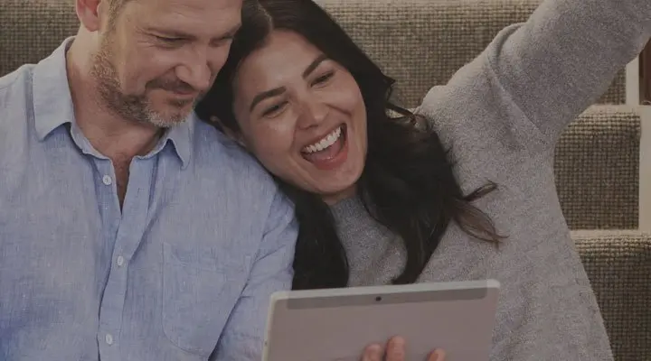 Pareja joven revisa contenido en una tablet mientras ríe sentada en un sofá.