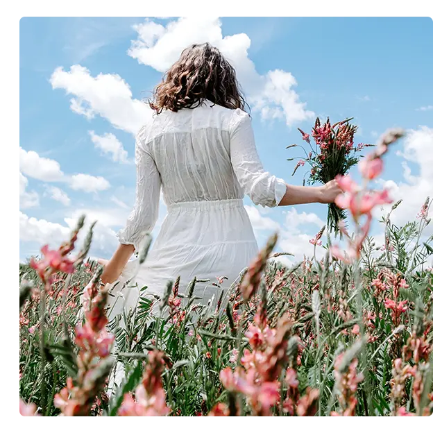 A woman in a white dress stands in a blooming wildflower field, holding a bouquet of pink flowers, with a blue sky and scattered clouds in the background.