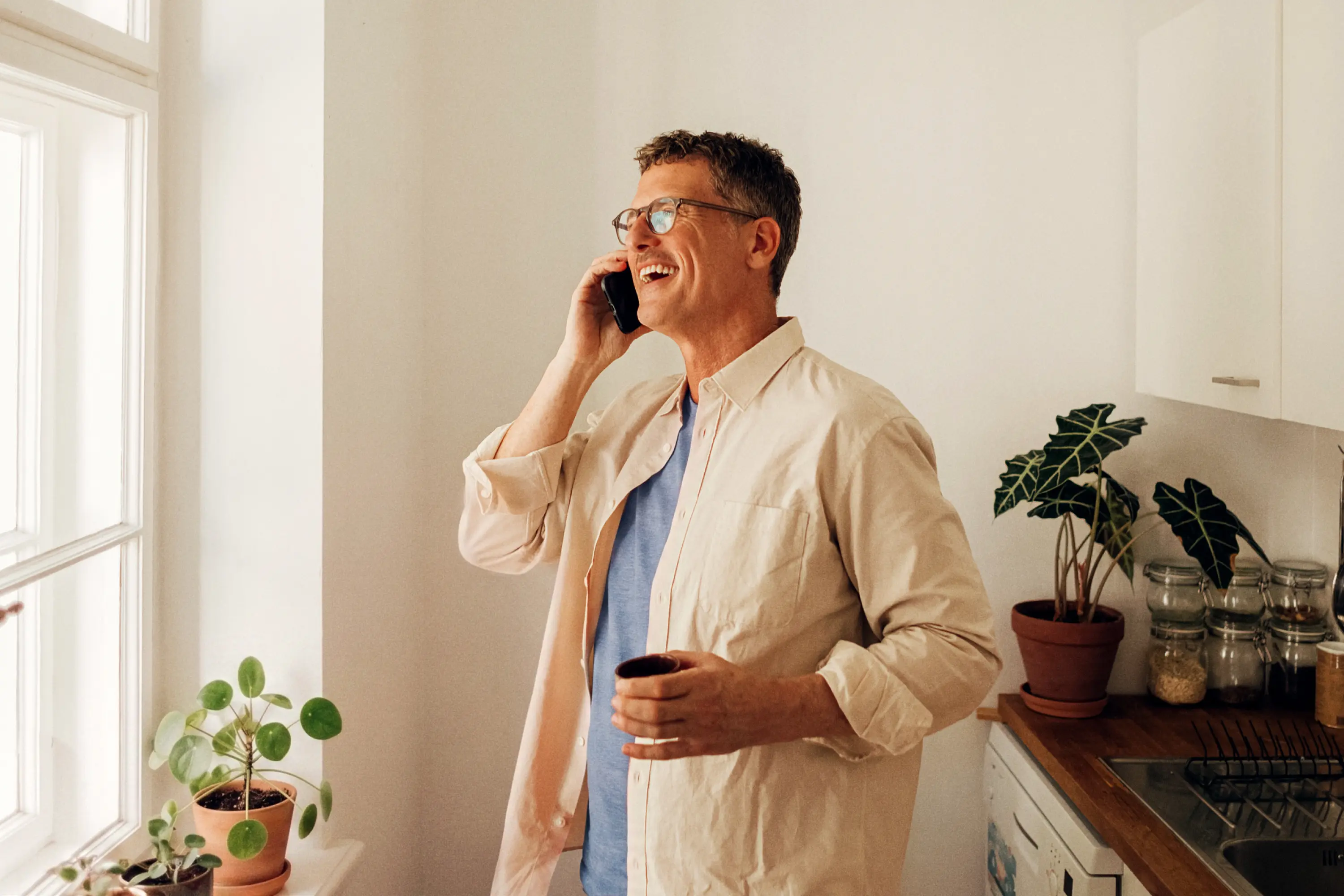 A middle-aged man with glasses laughing on a phone call while holding a mug in a bright kitchen near a window.