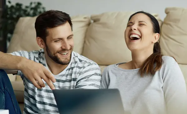 Mies ja nainen istuvat lattialla sohvan edessä tietokoneella ja nauravat.