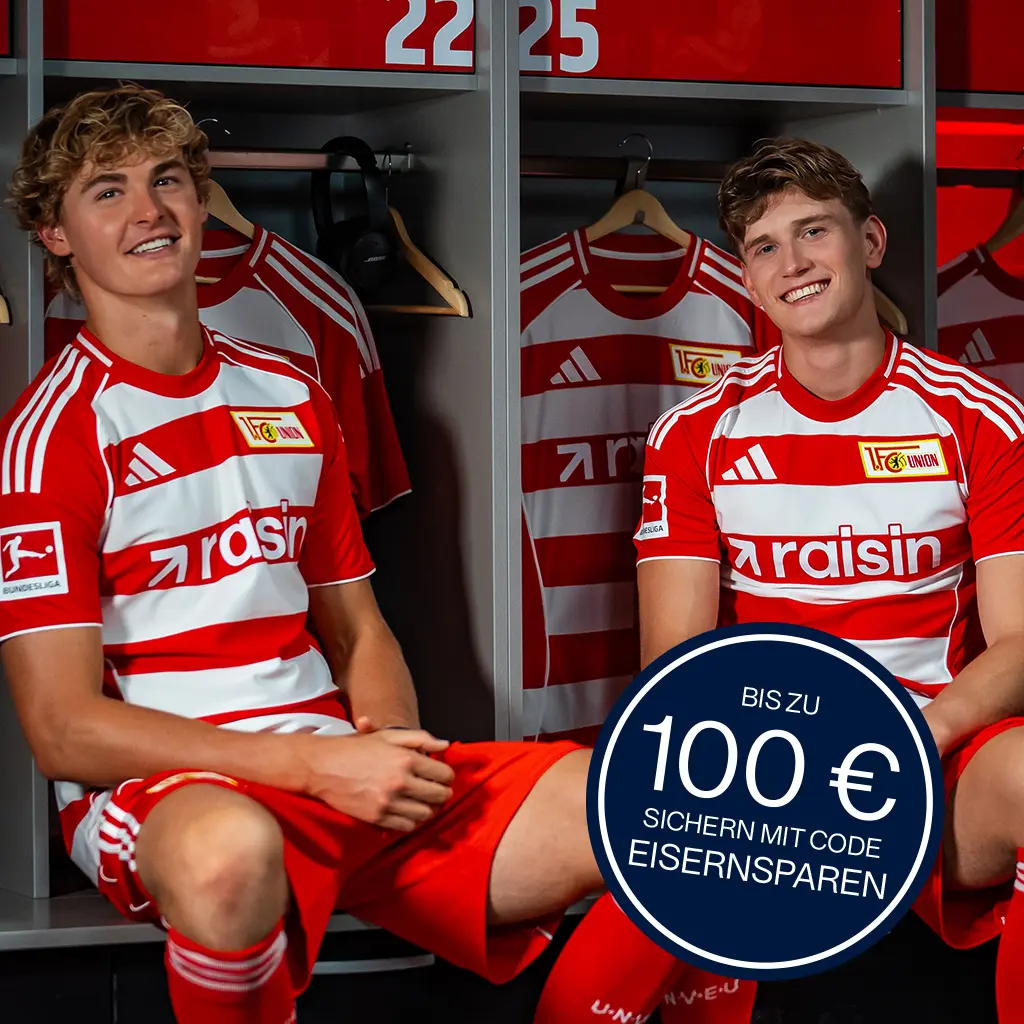Zwei Union-Berlin-Spieler sitzen in einer Umkleidekabine und tragen das Union-Berlin-Trikot mit dem Raisin-Logo auf der Brust, in der rechten Ecke ein Patch mit der Aufschrift "Bis zu 100 € sichern mit Code EISERNSPAREN"