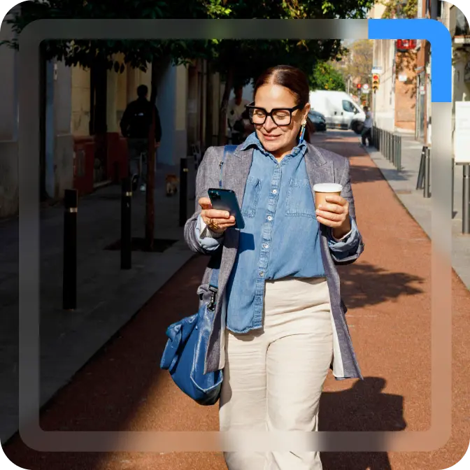 A woman in a denim shirt and grey jacket walks down a sunny street, while looking at her phone and holding a takeaway coffee cup