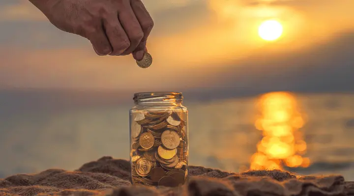 Tarro lleno de monedas con el mar de fondo y el sol en lo alto.