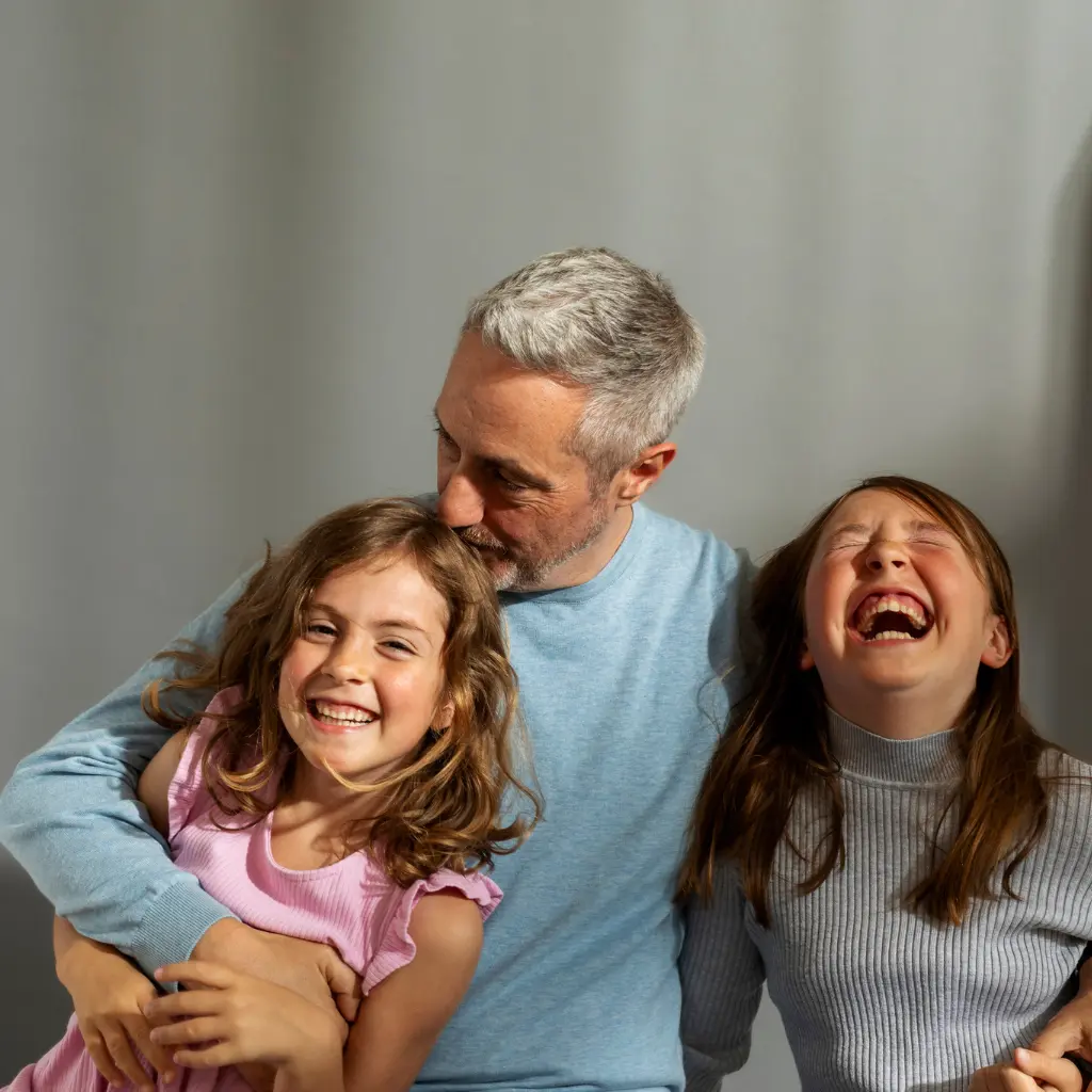 Ein Mann mit grauen Haaren umarmt zwei junge Mädchen, die herzlich lachen.