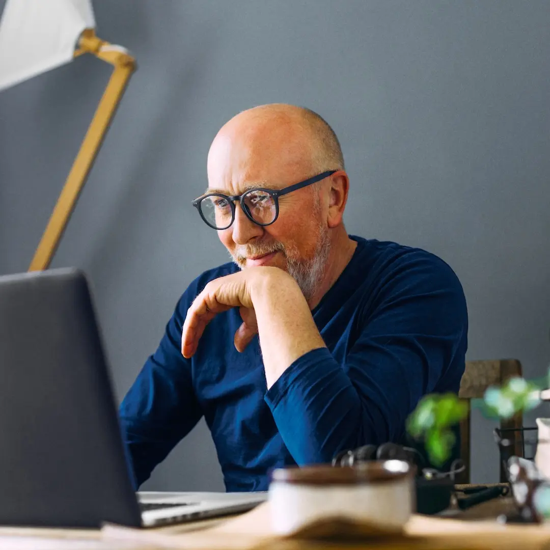 Middle-aged man with glasses looking thoughtfully at a laptop screen at his desk.