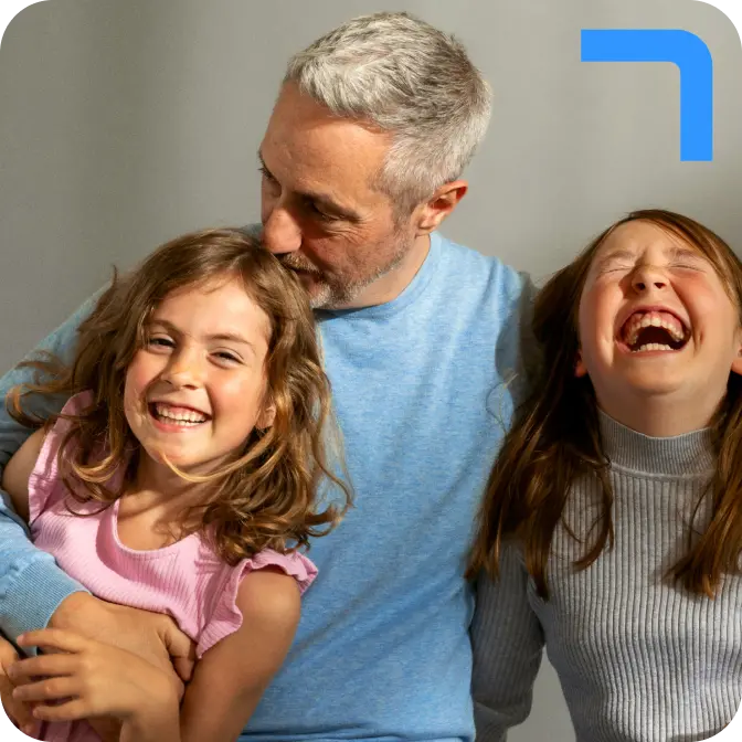 A grey-haired father wearing a long-sleeved light blue t-shirt hugs his two laughing young daughters, one wearing pink and the other wearing a grey turtleneck jumper.