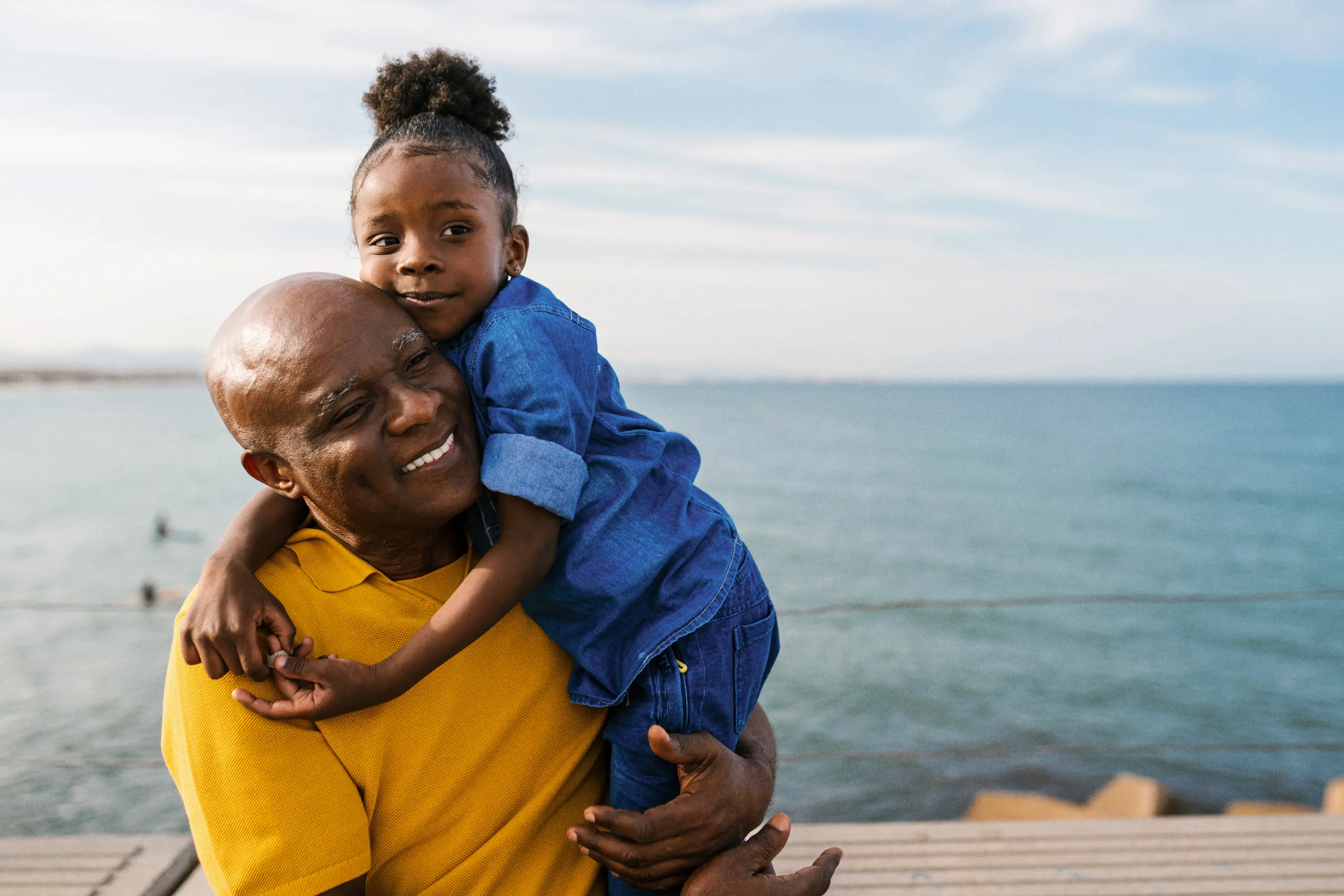 Raisin IE-Man and girl ocean A smiling older man in a yellow shirt holds a young girl hugging him, with the ocean in the background.