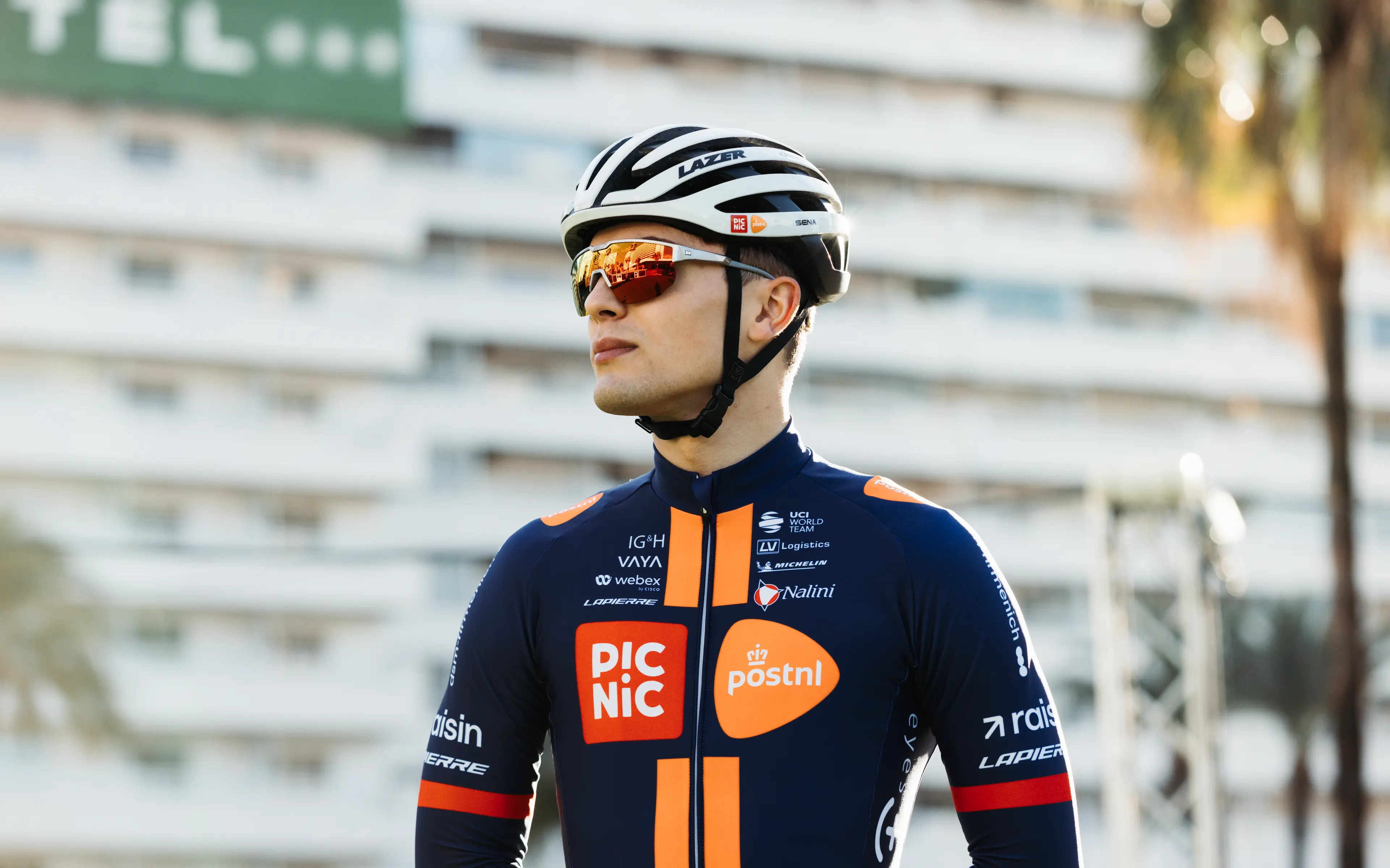 Professional cyclist wearing a Picnic PostNL jersey and helmet, standing outdoors with modern buildings blurred in the background.