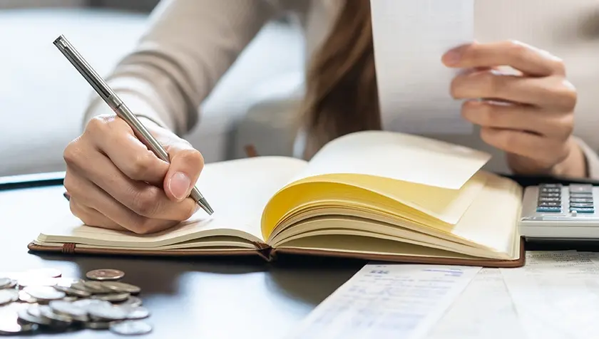 raisin_muistivihko_kynae_laskin.jpg Henkilö katsoo kuitteja ja kirjoittaa muistiinpanoja vihkoon. Vieressä pöydällä on laskin ja kasa kolikoita.