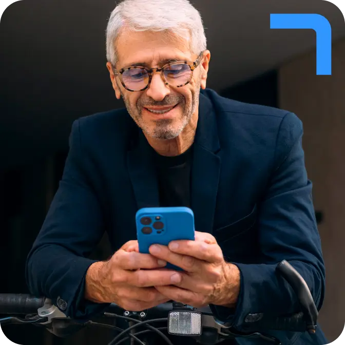 A smiling man with grey hair and glasses, wearing a blue shirt, leaning on a bike and looking at his phone