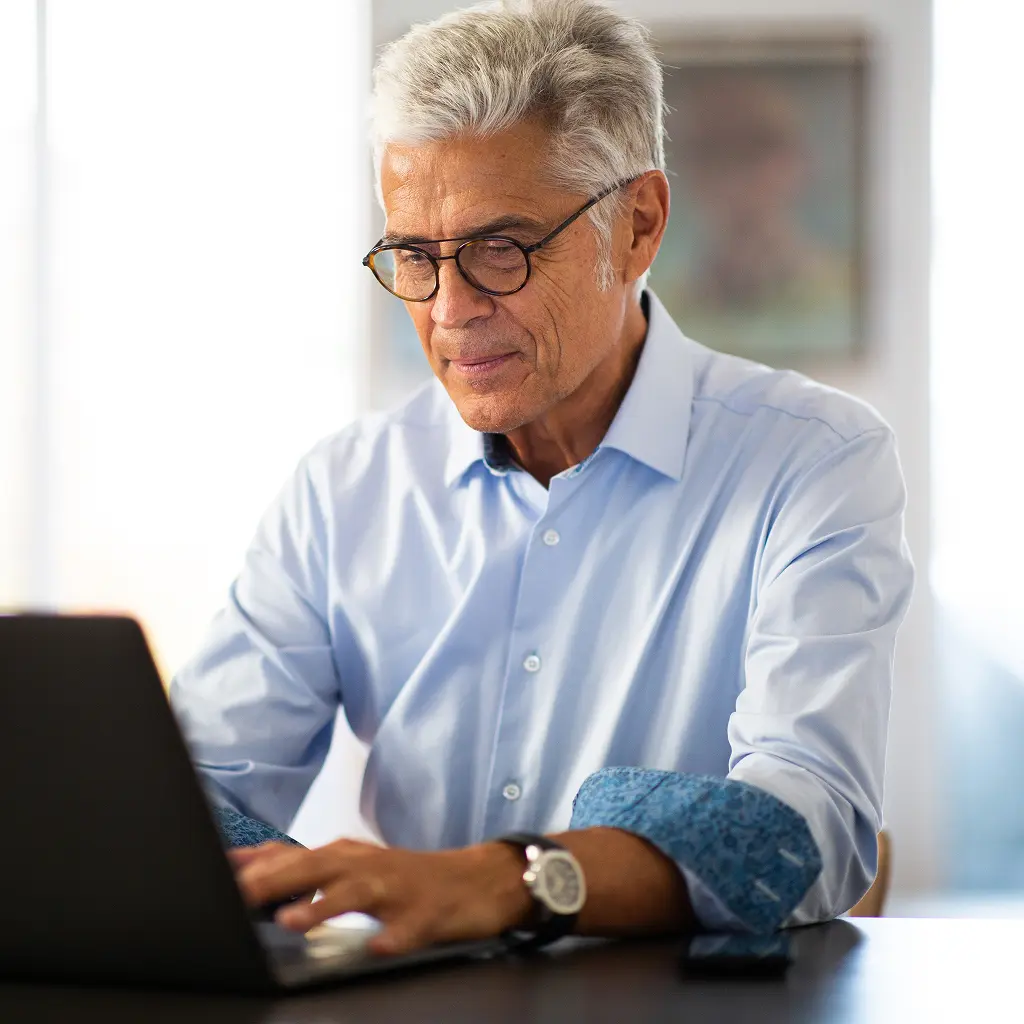 A senior man wearing glasses and a light blue button-up shirt sitting at a table working on a laptop.