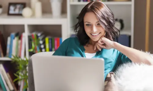Mujer cómoda en interior interactúa y sonríe frente a un portátil con estantería atrás.