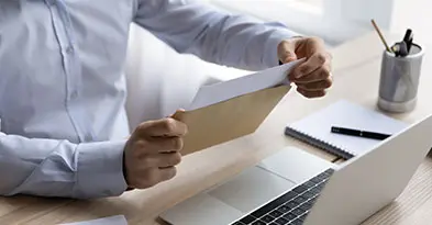 Raisin_FI_tietokone_kirje.jpg Henkilö avaa kirjekuorta työpöydän ääressä, jossa on kannettava tietokone, muistivihko ja kyniä.