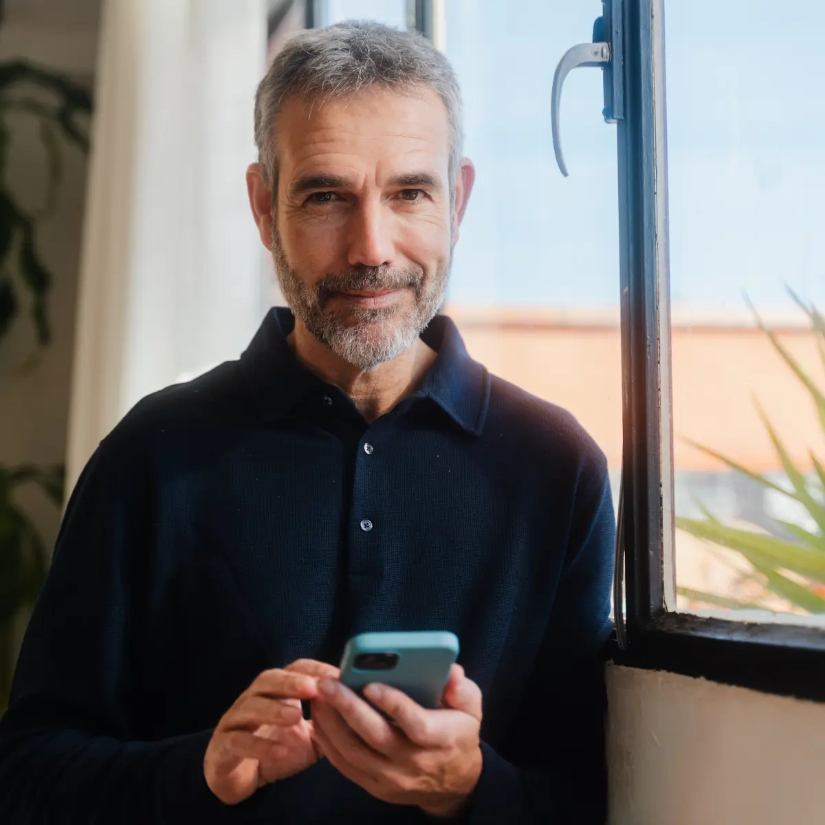 raisin_de_mann_smartphone_fenster_investment.jpg Mann blickt lächelnd in die Kamera, während er ein Smartphone vor einem Fenster hält.