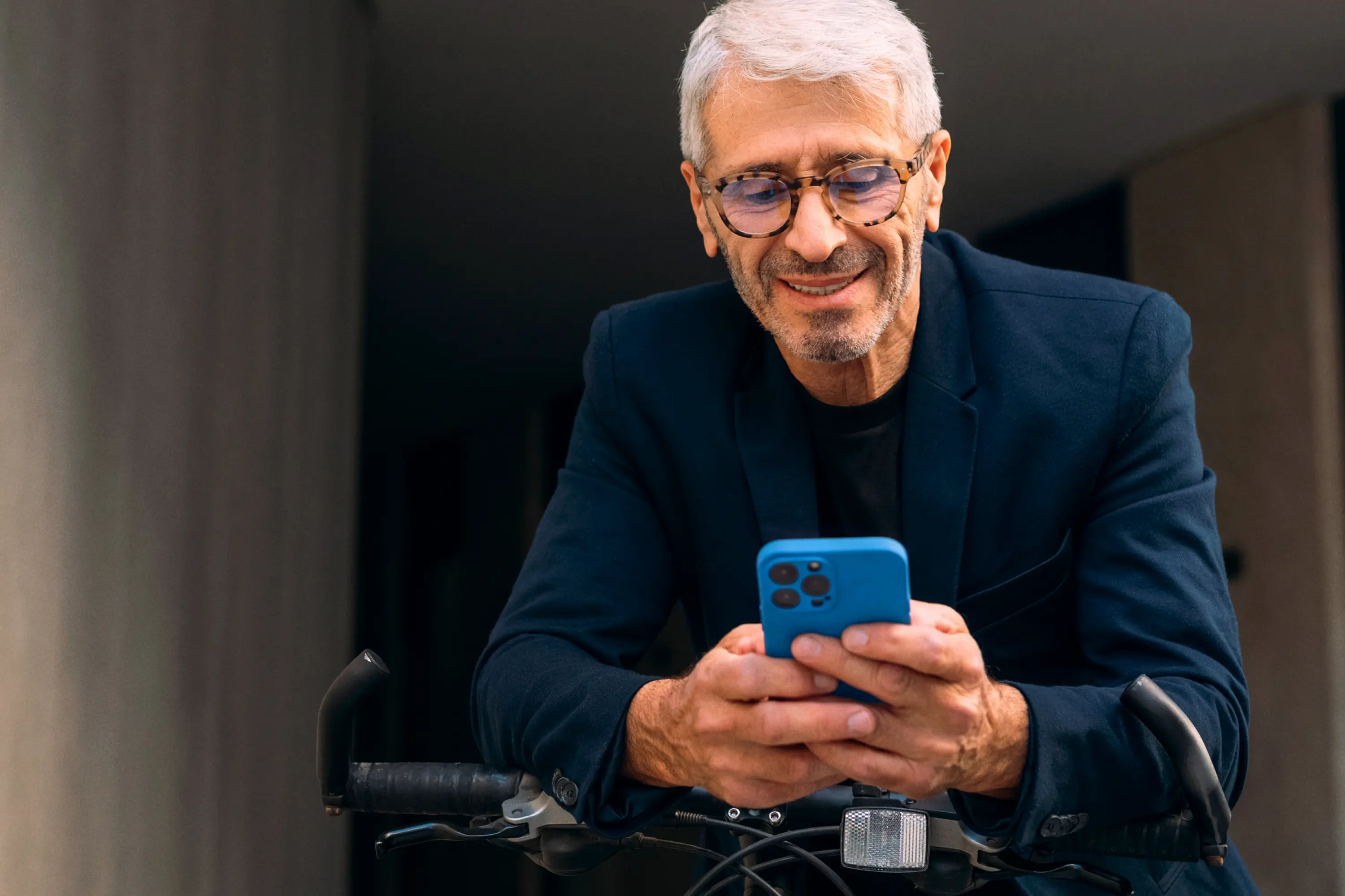 A smiling man with grey hair and glasses leaning on a bicycle while looking at his blue smartphone.