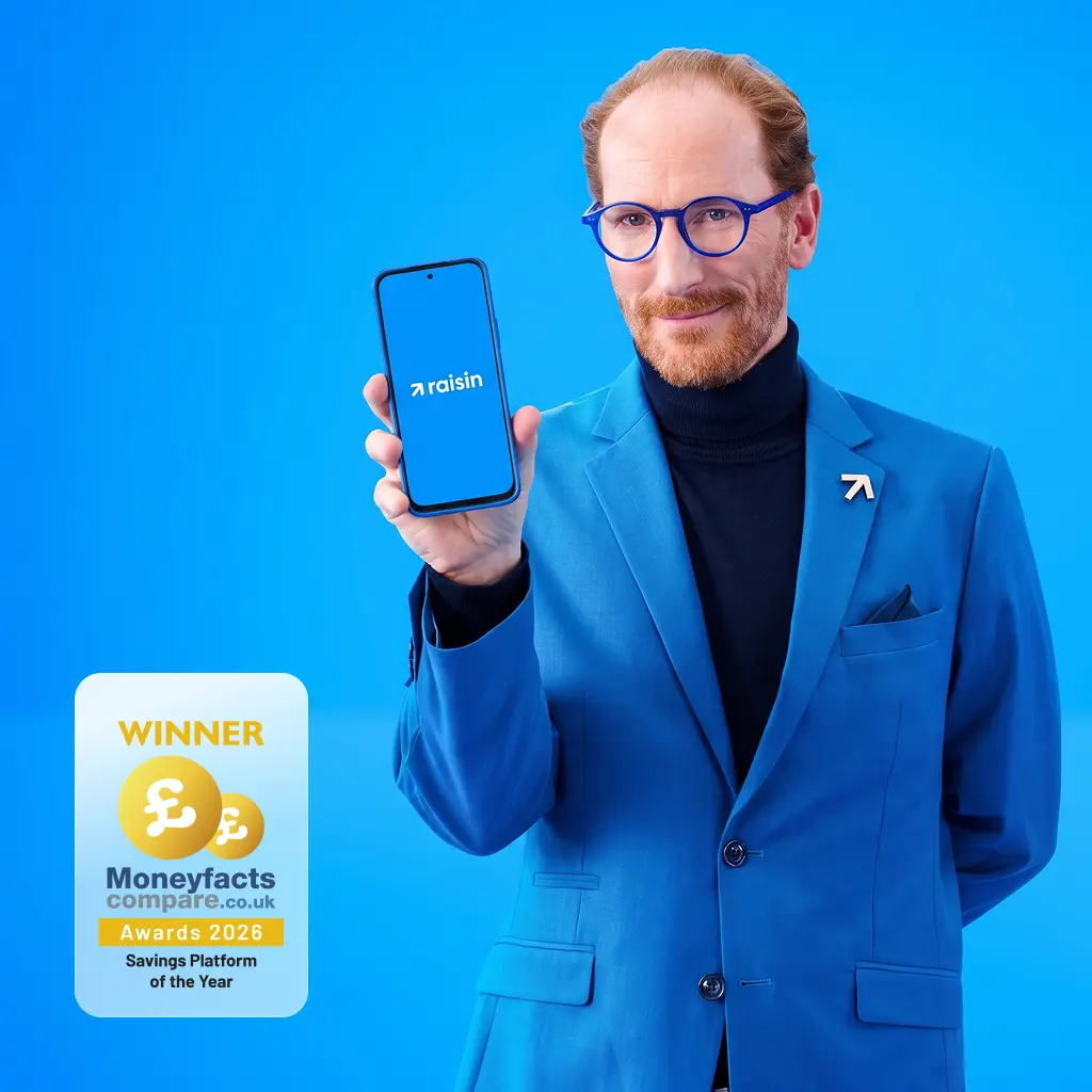 Man in a blue suit and glasses smiling while holding up a phone with the Raisin logo against a blue background.