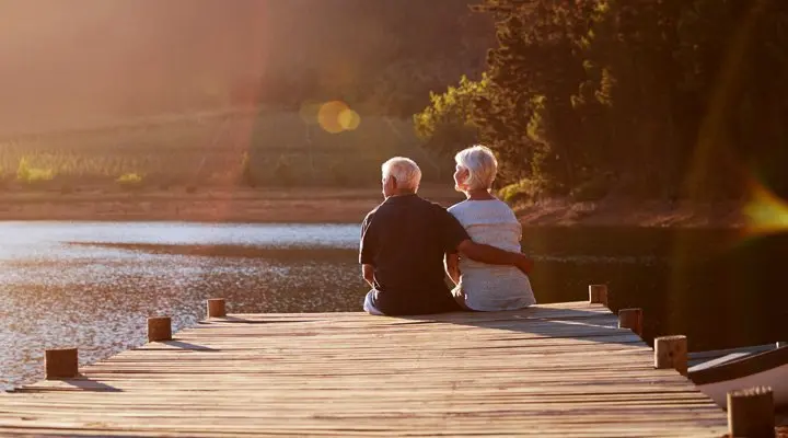Een ouder echtpaar zit hand in hand op een houten steiger en kijkt naar het water, wat ontspanning uitbeeldt.