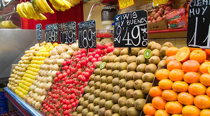 Puesto exterior exhibe manzanas, tomates y naranjas junto a carteles de precio.