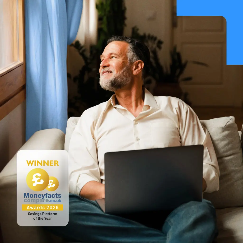 A smiling mature man sitting on a couch with a laptop, looking out a window. Features a Moneyfacts 2026 Savings Platform award badge.