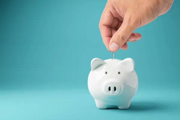 Een hand laat een munt vallen in een wit spaarvarken tegen een effen achtergrond, wat sparen en vermogensopbouw benadrukt.