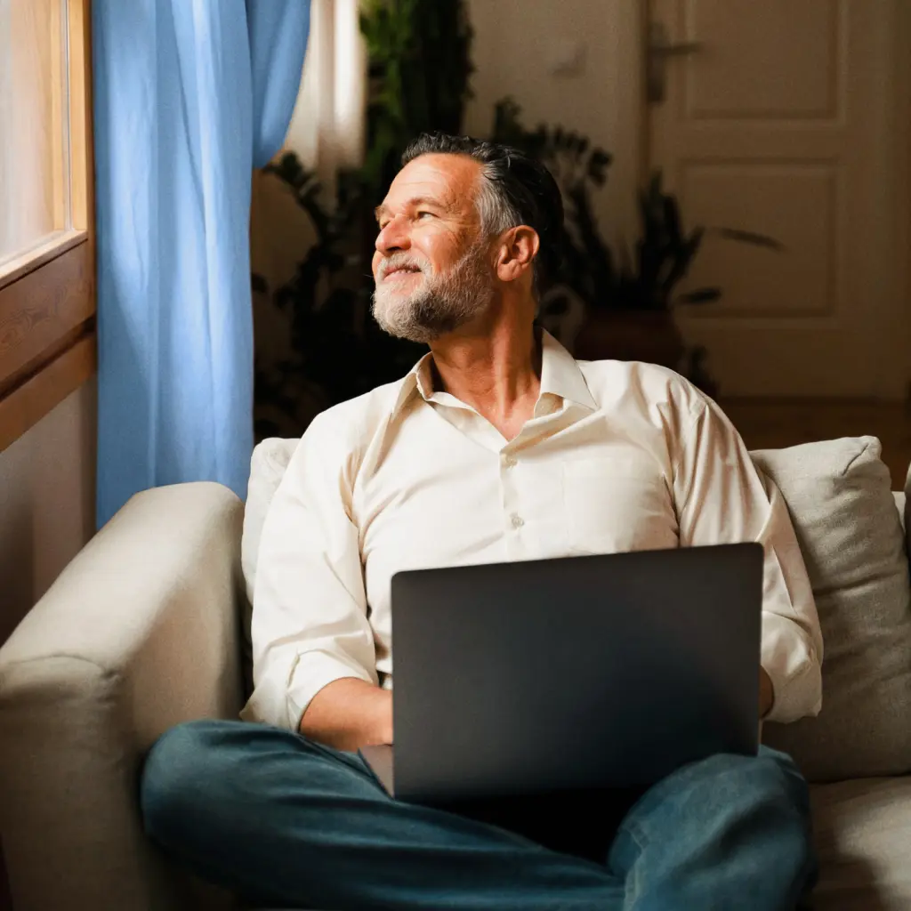 A man with a grey beard sitting on a sofa looking out of the window, with a laptop.