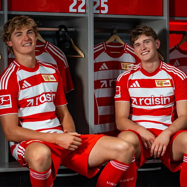 Zwei Fussballspieler des 1. FC Union Berlin sitzen in einer Umkleidekabine mit dem rot-weißen Raisin-Trikot