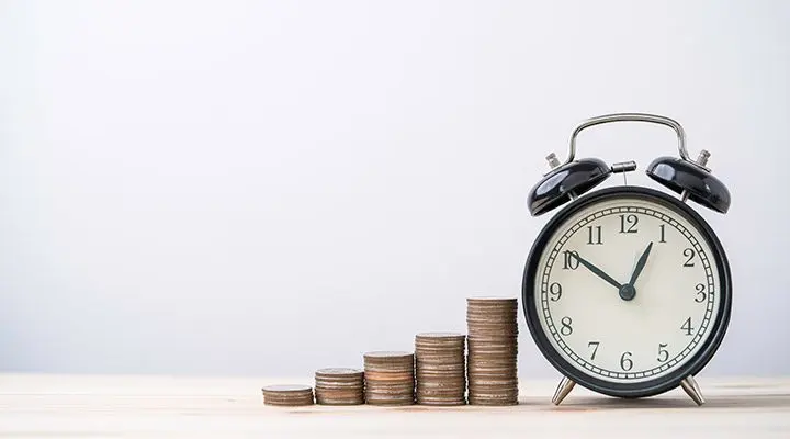 Despertador analógico acompaña pilas de monedas que aumentan en altura sobre la mesa.