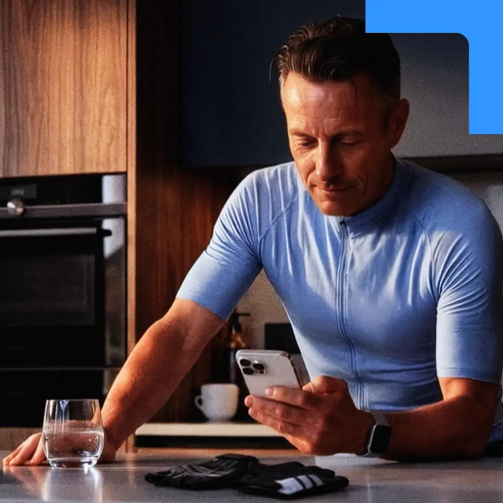 Een man in een lichtblauwe wielrennersoutfit leunt tegen een aanrecht in een keuken terwijl hij op zijn smartphone kijkt.