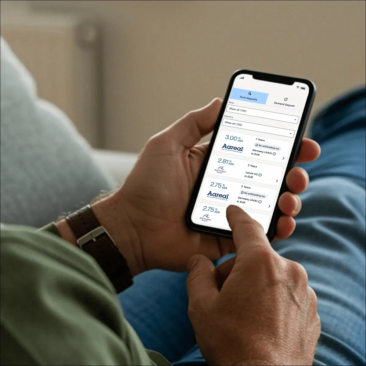 raisin_ie_how_to_register_step_2_smartphone Close-up of a man holding a smartphone while seated, browsing interest rate options on the Raisin app, with one finger poised to select an offer.