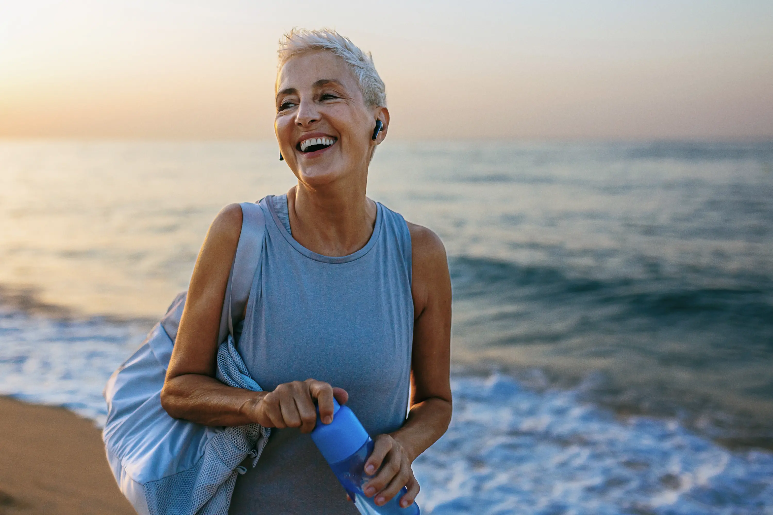 Raisin IE-Senior woman smiling on beach A smiling senior woman with short white hair walking along a beach, wearing activewear and holding a blue water bottle.
