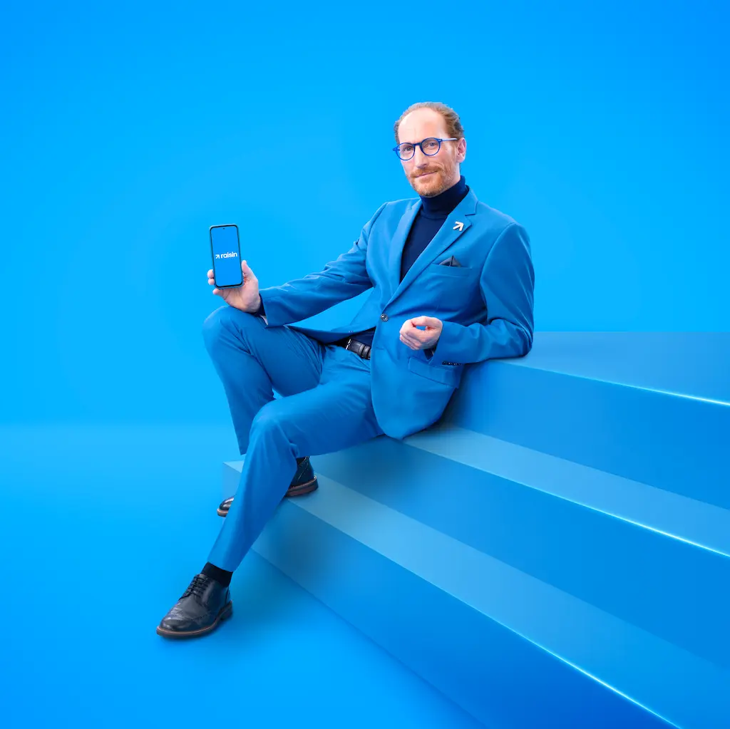 Spokesperson in a bright blue suit sitting on blue stairs holding a smartphone displaying the Raisin UK app.