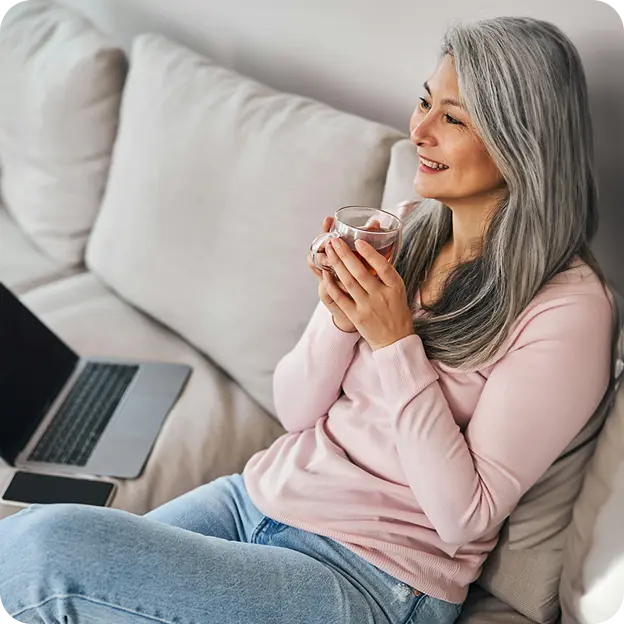 Nainen istuu sohvalla kahvikuppi kädessä ja tietokone vieressä