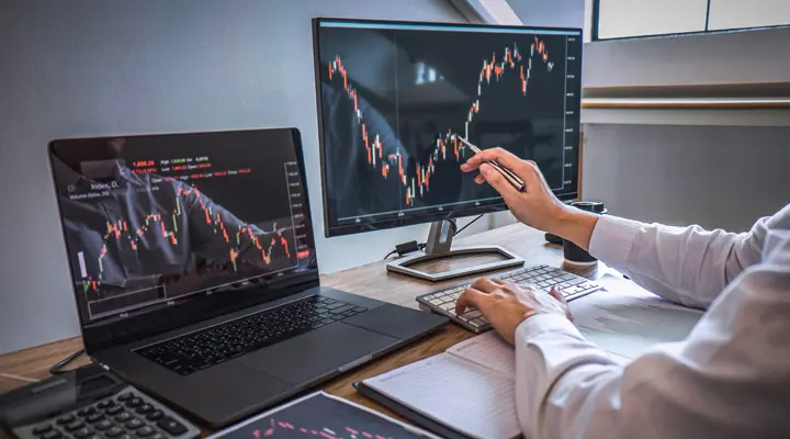 Een bureau met meerdere beeldschermen waarop financiële grafieken worden weergegeven, terwijl een hand ernaar wijst.