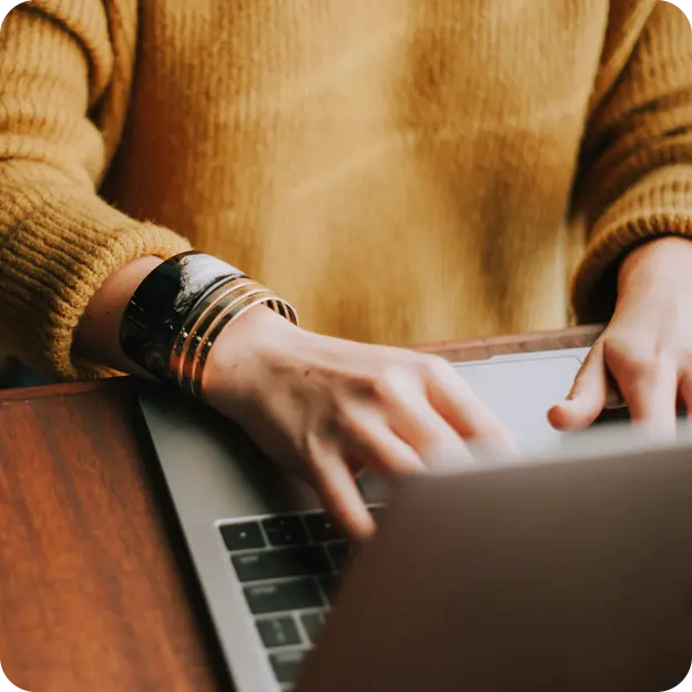 Person wearing a mustard-yellow sweater typing on a laptop at a wooden desk
