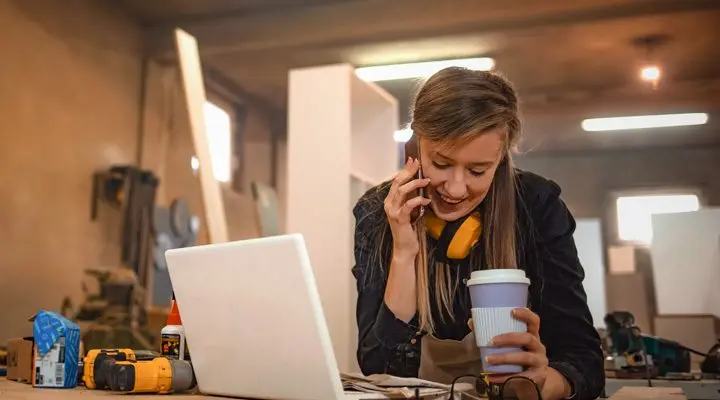 Een vrouw belt in een werkplaatsomgeving terwijl ze koffie vasthoudt en voor zich een laptop heeft, wat multitasken en zakelijk contact illustreert.