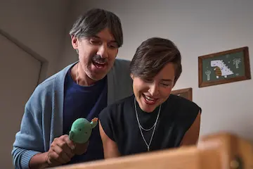 Zwei Erwachsene stehen nebeneinander und blicken lächelnd nach unten. Eine Person hält ein kleines grünes Stoffspielzeug in der Hand. Die Szene spielt in einem wohnlichen Innenraum und wirkt vertraut und verspielt.