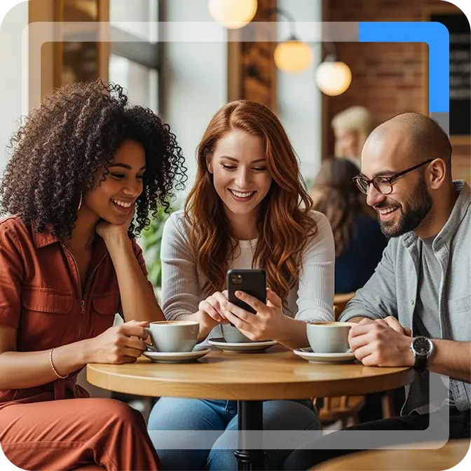 An image of three friends sitting around a round table drinking coffee. The person in the middle is showing the other two something on her phone.