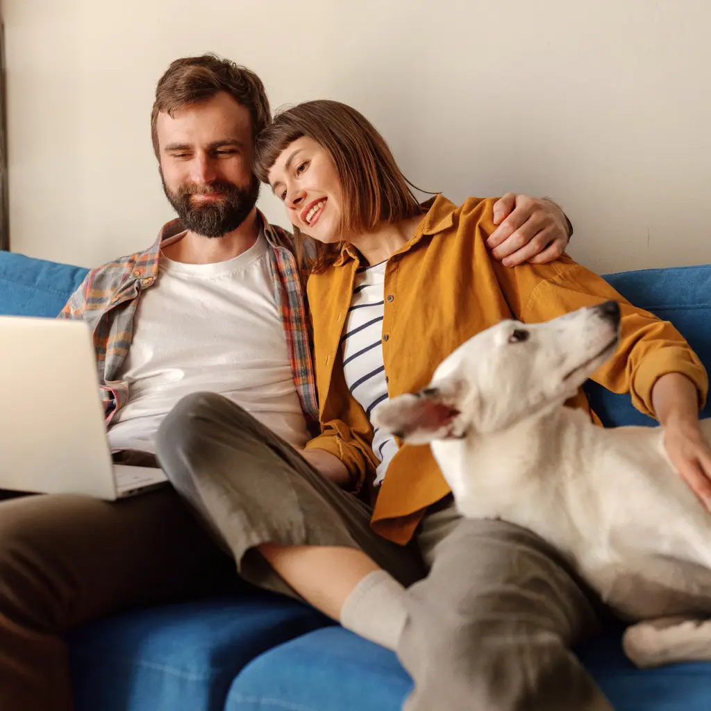 Un homme et une femme sont assis sur un canapé bleu en regardant un ordinateur portable, tandis que la femme caresse un chien blanc posé sur leurs genoux.