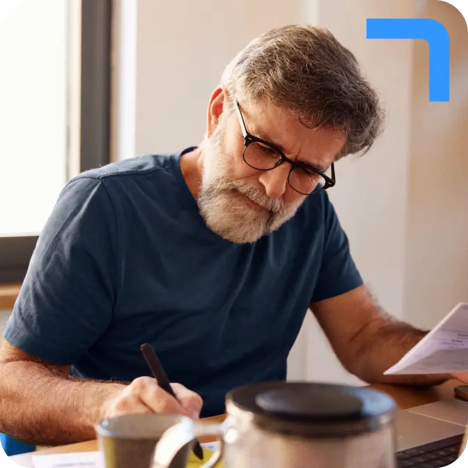 raisin-uk-hero-bearded-man-writing.png A bearded man in a blue t-shirt and glasses writes using a pen. He holds a piece of paper in his other hand.