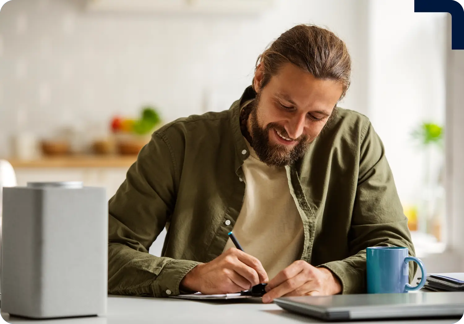 Man with long hair and beard writing in notebook at home, smiling. Laptop and coffee mug on desk. Bright, cozy setting with natural light.