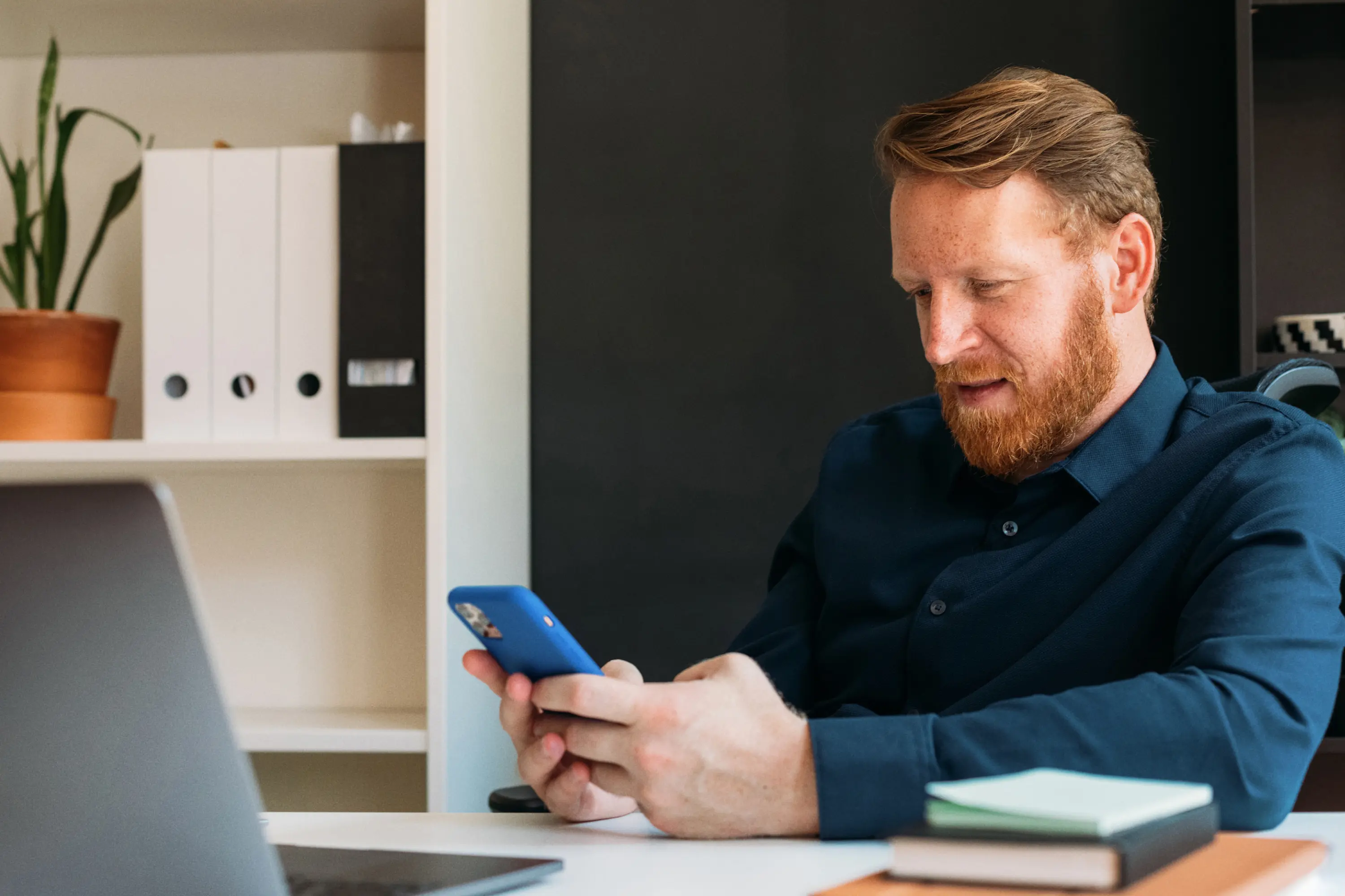 A man with a red beard sitting at a desk with a laptop, looking closely at his smartphone in a blue case.