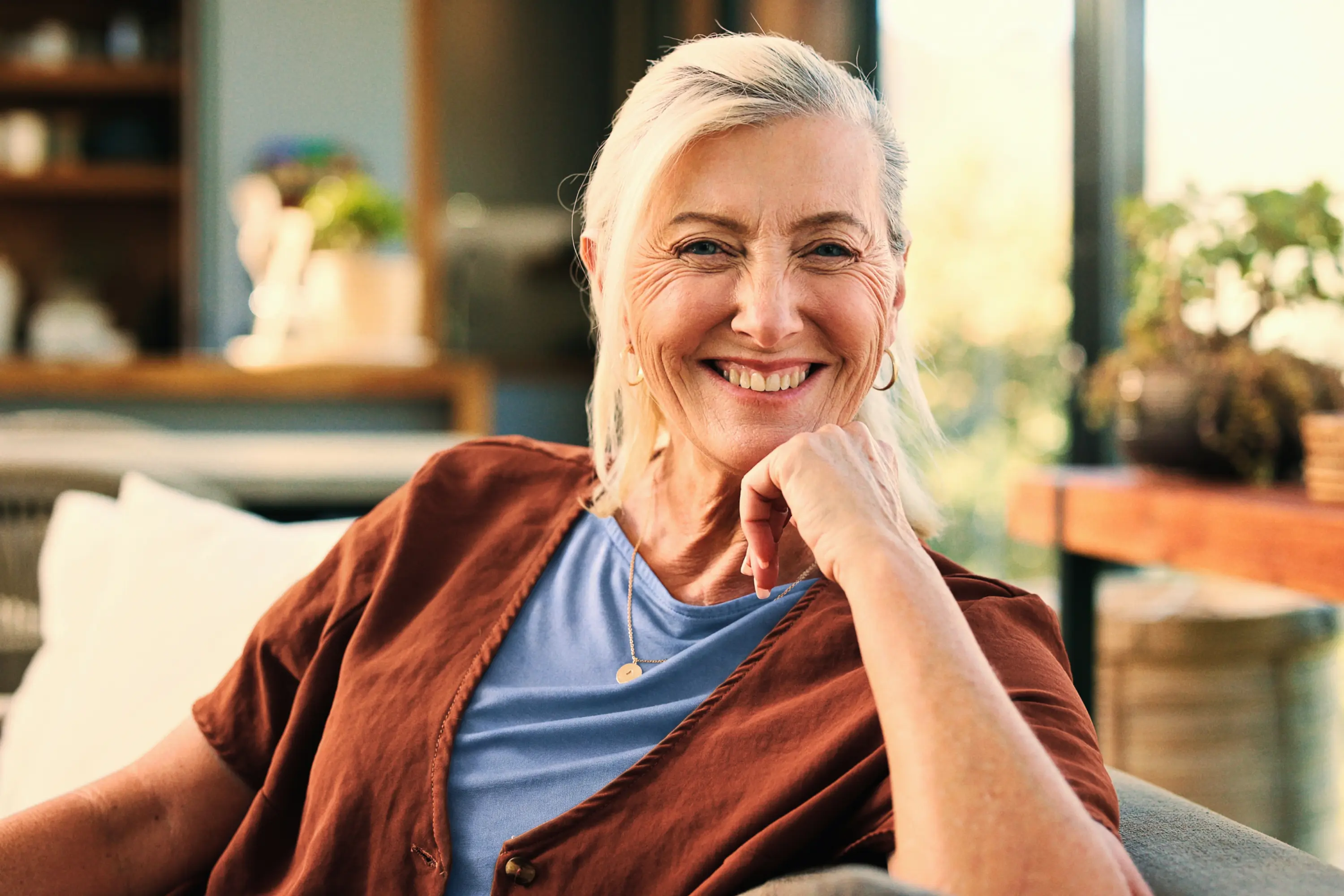 A smiling senior woman with blonde hair resting her chin on her hand, wearing a blue shirt and rust-coloured cardigan.