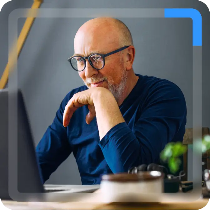An older man with glasses sitting at a desk, looking attentively at a laptop screen.